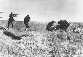 Photo noir et blanc prise à Ivangorod, en Ukraine. Dans un champ herbeux, sous un ciel clair, un soldat allemand (à gauche, au second plan), de profil, jambes écartées, tient en joue une femme qui lui tourne le dos, un enfant dans les bras. Devant le soldat, on distingue un corps de femme étendu sur le sol. Sur le bord gauche de la photo, deux canons de fusils, l’un au-dessus de l’autre, sont aussi visibles. Et, sur la droite, quatre hommes semblent creuser un trou.