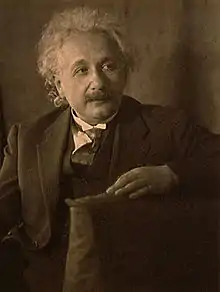 Photographie sépia d'un homme assis au cheveux gris clair, en coiffure désordonnée, portant une moustache, ayant le regard pensif tourné vers la gauche.