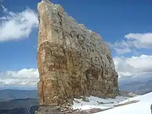 Une immense barrière verticale de roc se dresse, un peu penchée, dans le ciel. Au bas, un homme ressemble à une fourmi.