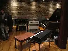 photo d'un piano et de claviers dans une pièce au mur de briques et au sol parquet.