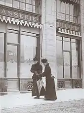 Photographie en noir et blanc de deux dames habillées sombrement. La première se tient droite et la seconde est légèrement penchée.