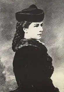 Portrait en noir et blanc à mi-corps de profil droit de Sissi portant une toque en fourrure de laquelle s'échappent de longs cheveux tressés.