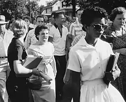 Photo en noir et blanc d'une jeune fille noire, les livres sur le bras, poursuivie par une femme blanche la bouche grande ouverte.