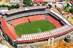 Photographie en couleurs. Vue aérienne d'un stade aux tribunes et aux toits couleur terre, entouré de son milieu urbain.
