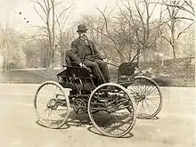 Photographie en noir et blanc. Un homme est assis sur le siège d'une automobile.