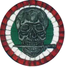 Cercle de plusieurs couleurs avec une tête de mort tenant un couteau entre ses dents.