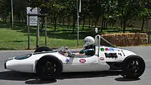 Photographie en couleur d'une monoplace de Formule Junior blanche, vue de profil, sur une route.