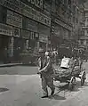 Passant devant un immeuble presque recouvert de panneaux publicitaires, jeune apprenti peintre en bâtiment tirant un chariot.