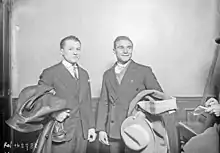 Deux hommes en costumes, côte à côte, tiennent leurs manteaux à la main.