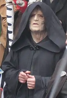 Homme qui porte un masque de vieillard et une robe à capuchon noir.