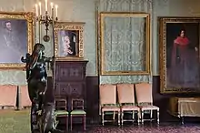 Photo de l'intérieur d'un musée comprenant diverses statues, peintures et chaises. Au milieu se trouve un grand cadre vide, laissant apparaître la tapisserie verte-bleue du mur.