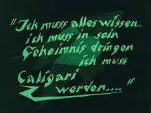 Un écran noir avec des formes vertes délavées en arrière-plan, et des mots verts en langue allemande écrits en caractères d'imprimerie angulaires au premier plan.