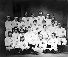Photographie en noir et blanc représentant un groupe d'hommes posant debout ou assis dans un espace fermé sombre. Ils sont tout de blanc vêtus.