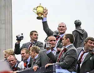 Les joueurs anglais sont en costume et font la fête, montrant le trophée de la Coupe du monde à leurs supporters.
