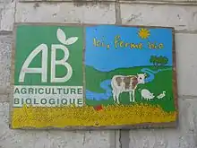 C'est une pancarte avec une peinture simplifiée d'un élevage (une vache, un cochon, une poule) avec le titre : "Une ferme bio". Les couleurs sont le bleu, le jaune et le vert. On retrouve aussi le logo AB.
