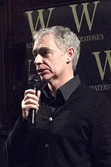  Eoin Colfer,en costume noir avec un microphone à la bouche, certainement en conférence de presse