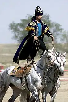 Kazakh en costume traditionnel, debout sur deux chevaux en mouvement.