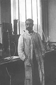 Photographie d'un homme en blouse blanche, debout appuyé sur une paillasse de laboratoire.