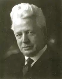 Photographie du buste d'un homme en noir et blanc. L'homme aux cheveux blancs porte un costume et une cravate noirs. La tête penchée à gauche il observe le spectateur.