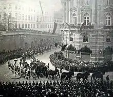 Photo en noir et blanc montrant un cercueil tiré par quatre paires de chevaux empruntant une rue courbe et entouré par des forces de l'ordre qui contiennent une foule assez dense.
