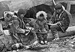Une famille Inuit est assise sur un tronc devant une tente. Les parents, portant de chauds vêtements fait de peaux animales, s'occupent de tâches ménagères. Entre eux est assis un jeune enfant, habillé de la même façon, qui regarde l'appareil photo. Un bébé est suspendu au dos de la mère.
