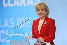 Photographie d'une femme blonde, debout, prononçant un discours, portant une veste rouge.