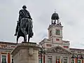 Monument du roi Charles III au centre de la place, devant la Maison royale du courrier et son horloge.