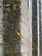 Photographie en couleurs d'un mur dont une pierre en forme d'équerre est signalée par une flèche jaune.