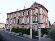 Maison ancienne avec briques rouges en façade, sur trois niveaux, mur de clôture, portail et haie.