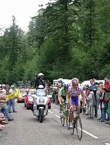 Coureur avec maillot blanc à pois rouge de meilleur grimpeur en danseuse devant un coureur en maillot vert et bleu, encadrés par des haies de spectateurs.