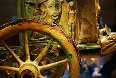 Photographie montrant une roue à rayons en bronze oxydé et en arrière plan le corps même du char avec des bas-reliefs.