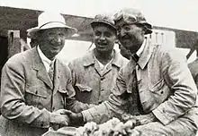 Photo de trois hommes, l'un en tenue de pilote reçoit les félicitations d'un autre en costume, sous le regard d'un mécanicien