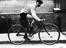 Photographie en noir et blanc d'un cycliste sur sa bicyclette, les mains tenant le guidon, la jambe gauche à terre