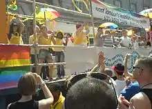 Des gens regardent des femmes vêtues en jaune dansant sur un char, sur lequel un drapeau arc-en-ciel est accroché