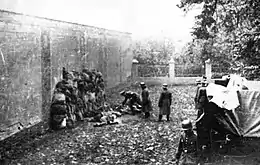 Photographie en noir et blanc de civils debout, adossés le long d'un mur à gauche, alors que des membres d'un groupe d'intervention, à droite, les fusillent.