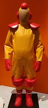 Photo en couleur d'un uniforme jaune et rouge à l'allure bizarre et d'une affiche du film La Soupe aux choux encadrée au mur.