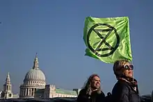 Photo couleur d'une femme de profil portant, sous un ciel bleu, un drapeau au fond vert, frappé d'un cercle noir contenant un sablier figuré par deux triangles accolés.