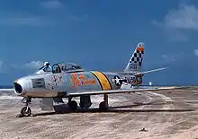 L'avion de chasse argenté de Glenn sur le tarmac, avec une bande jaune derrière le cockpit et un motif en damier sur la queue.