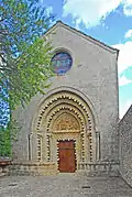 Façade étroite et claire, percée d'un œil-de-bœuf et d'un portail arrondi. Un arbre au feuillage vert clair le masque légèrement, à gauche.