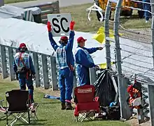 Des commissaires de course agitent simultanément un drapeau jaune et le panneau indiquant l'entrée de la voiture de sécurité en piste.