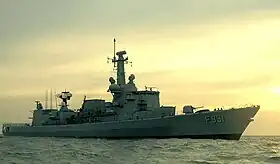 Photo d'un navire militaire de teinte grise voguant sur une mer calme sous un ciel rosâtre.