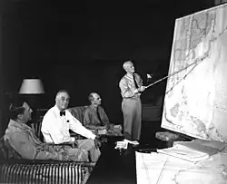 Trois hommes sont assis dans des fauteuils. Un autre est debout et tient une baguette indiquant le Japon sur une carte murale du Pacifique.