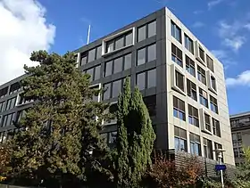 Photo d'un bâtiment gris rectangulaire, caché derrière un arbre, sur lequel certaines fenêtres présentent une mise en relief.