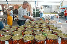 Sous un chapiteau, une quarantaine de grandes boîtes de haricots verts en morceaux sont ouvertes et prêtes à être cuisinées ; deux personnes, gantées, s’en occupent.