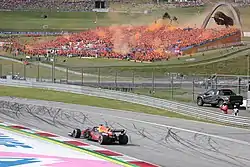 A matte navy single-seater open-wheel racing drives around a corner on a tarmac course while a grandstand full of people dressed in orange set off orange fireworks in the background.