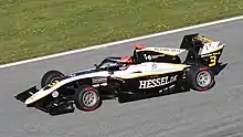 Photographie d'une monoplace de Formule 3 blanche et noire avec des traits jaunes, vue de trois-quarts gauche, d'en haut, sur une piste sèche.