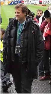 un homme debout muni d'une accréditation, est vêtu d'un manteau, dans une tribune près d'un terrain de rugby.
