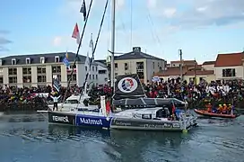 Bateau noir, bleu et gris. Le navigateur, de dos, salue des deux bras la foule massée sur le quai.