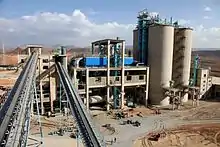 vue d'une usine avec deux convoyeurs au premier plan, un bâtiment de type industriel et quatre silos à leur droite