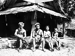 Photo en noir et blanc figurant une famille assise devant sa maison en bois.